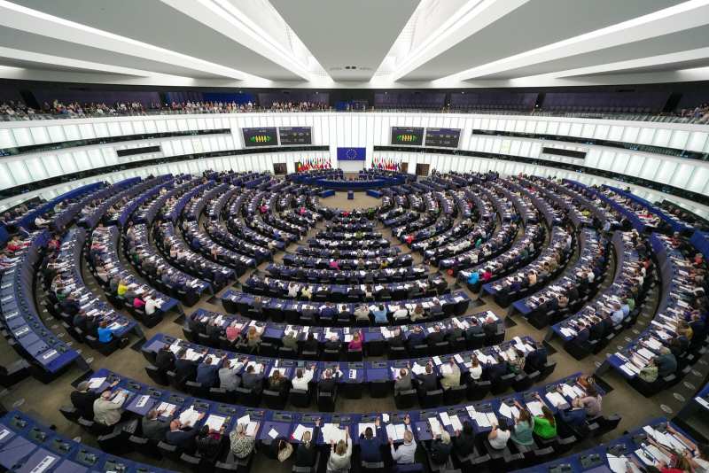 Vue plongeante en grand angle sur un vaste hémicycle aux pupitres bleus disposés en arcs concentriques, occupés par des centaines de personnes ; au fond, la tribune, un drapeau européen et deux rangées de drapeaux nationaux, sous un plafond blanc à lames lumineuses.