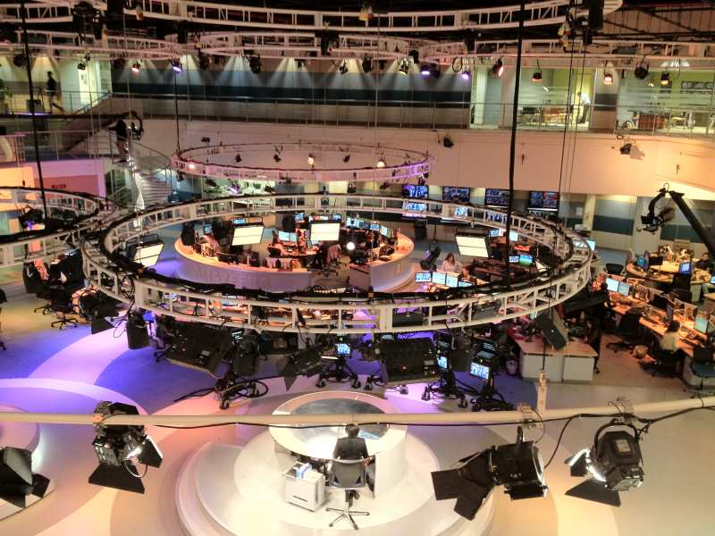 Vue plongeante sur un vaste plateau de télévision circulaire entouré de postes de travail et d'écrans, sous des rampes de projecteurs.