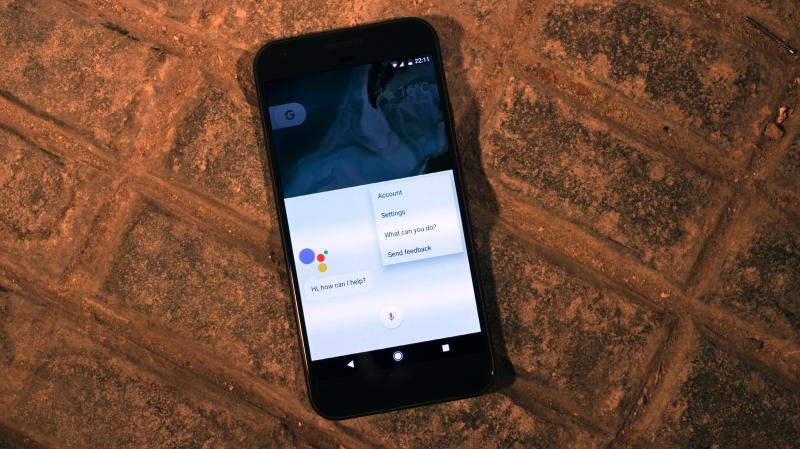 Un smartphone Android posé sur des pavés : en haut de l'écran d'accueil, le bouton de recherche Google ; en dessous, l'Assistant Google ouvert affiche « Hi, how can I help? ».
