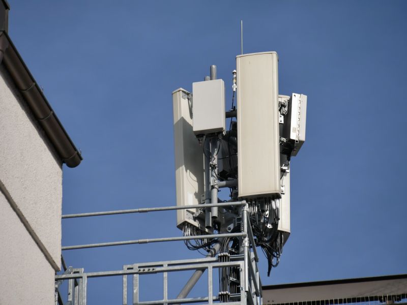 Vue en contre-plongée d'un mât d'antennes relais sur un toit : de longs panneaux d'antennes et de petits boîtiers d'antennes 5G fixés à une structure métallique.