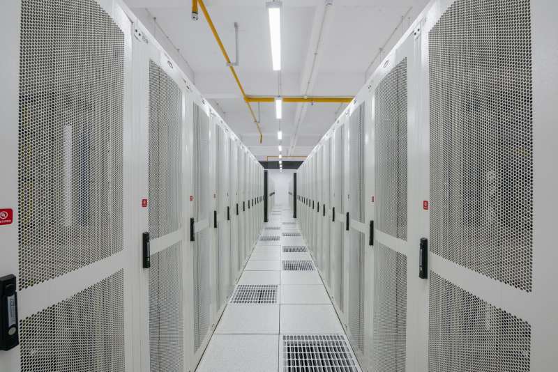 Une allée de centre de données entre deux rangées de baies de serveurs blanches aux portes perforées, sous une file de luminaires au plafond.