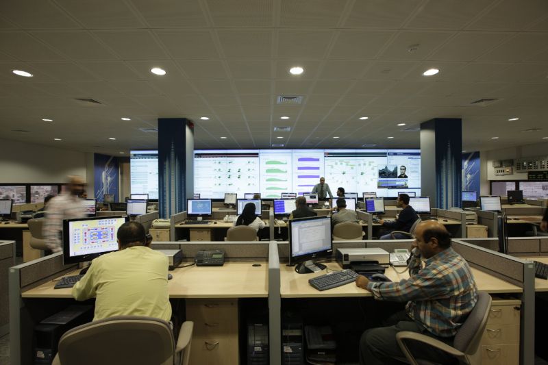 Une salle de supervision informatique : des techniciens travaillent à des postes à deux écrans, face à un mur d'écrans affichant graphiques et cartes de réseau.