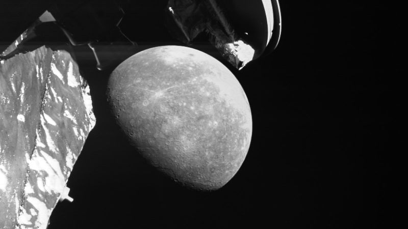 Photo en noir et blanc prise par une caméra de surveillance de BepiColombo : Mercure, à moitié éclairée et criblée de cratères, apparaît sur fond noir, encadrée par des éléments de la sonde au premier plan, dont l’arrière d’une antenne en haut.