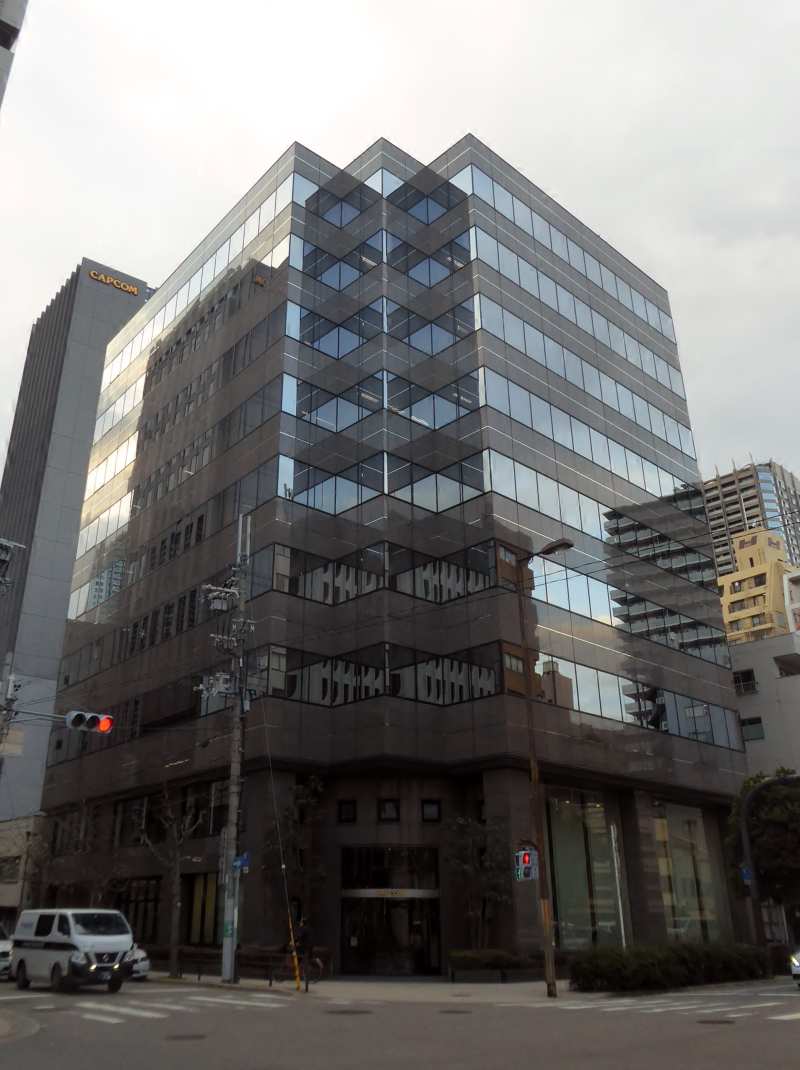 Carrefour d'Osaka bordé d'immeubles de bureaux vitrés ; au fond à gauche, une tour porte l'enseigne Capcom.