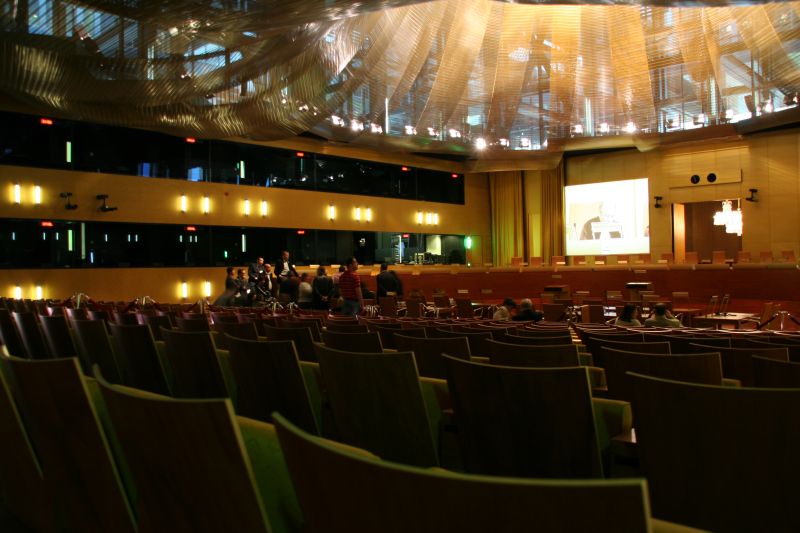 Grande salle d'audience vue depuis les rangs du public : fauteuils en bois au premier plan, bancs des juges et écran au fond, plafond tendu de voiles dorés ; quelques visiteurs debout.
