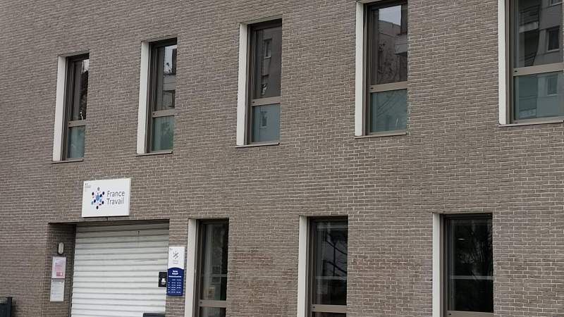 Façade de brique gris-brun percée de hautes fenêtres sur deux niveaux ; au rez-de-chaussée, à gauche, une enseigne blanche « France Travail » au-dessus d'un rideau métallique baissé, et une plaque bleue d'horaires d'ouverture à côté de l'entrée.