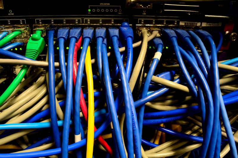 Gros plan sur un commutateur réseau : des dizaines de câbles Ethernet bleus, gris, rouge, jaune et vert branchés sur ses prises, voyants verts allumés.