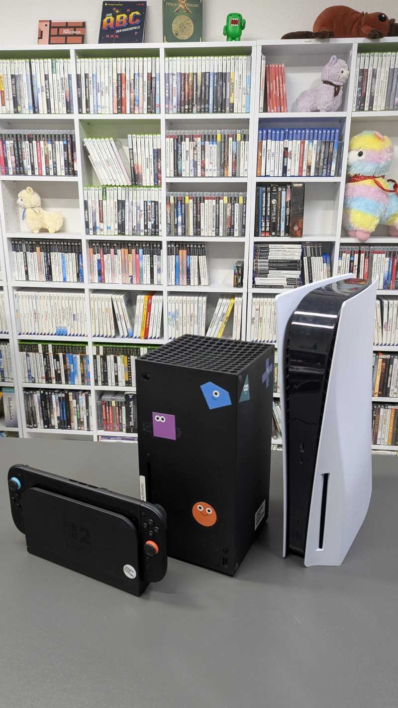 Une Nintendo Switch 2 insérée dans sa station d'accueil, une Xbox Series X noire ornée d'autocollants colorés et une PlayStation 5 blanche posées côte à côte sur une table grise, devant des étagères remplies de boîtes de jeux.
