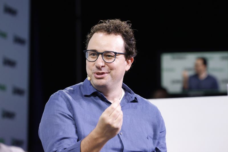 Dario Amodei, en chemise bleue et lunettes, parle sur scène, un micro à l'oreille, devant un fond sombre où un écran le montre en retransmission.