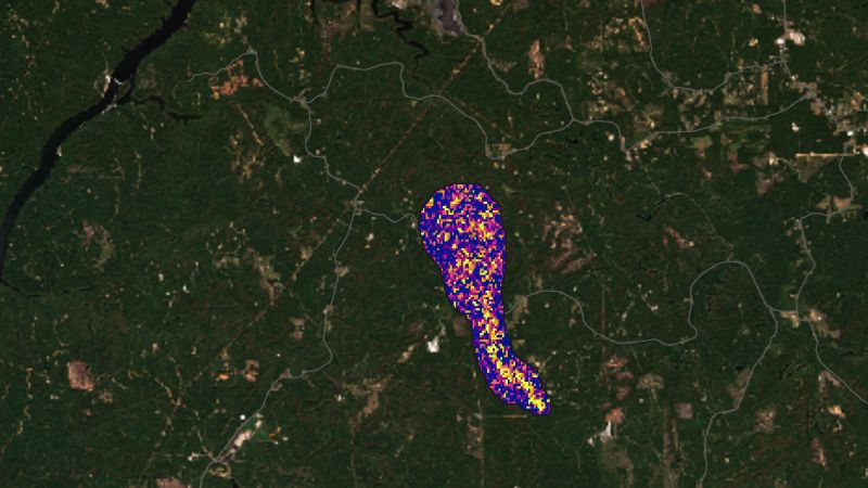 Vue satellite d'une région boisée vert sombre, traversée à gauche par une rivière sinueuse et par quelques routes grises ; au centre, une traînée allongée de pixels violets, roses et jaunes, orientée du haut vers le bas à droite, marque la concentration de méthane.