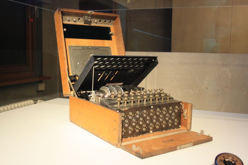 Machine Enigma dans sa caisse en bois ouverte, sous vitrine : plaque d'instructions en allemand dans le couvercle, capot intérieur relevé sur les rotors, clavier et, à l'avant, tableau de fiches.