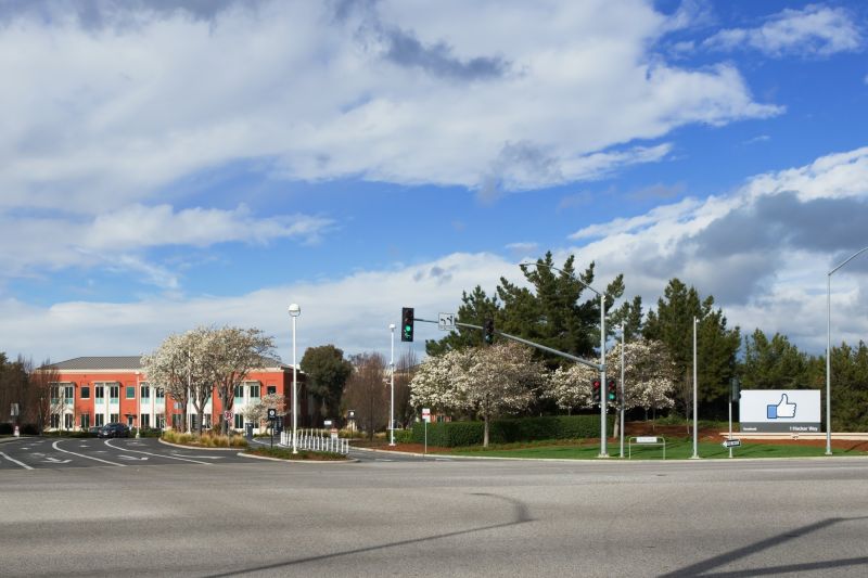 Un carrefour devant le complexe de Facebook à Menlo Park : un bâtiment rouge à deux étages, des arbres en fleurs et, à droite, le panneau au pouce levé du 1 Hacker Way.