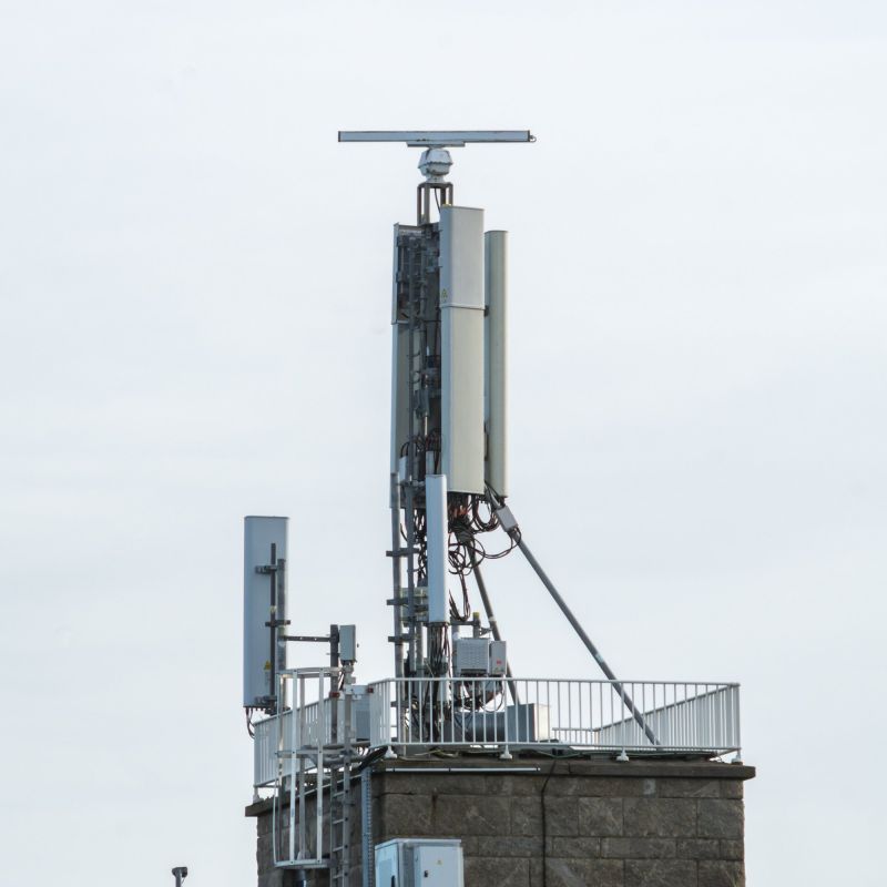 Mât d'antennes sur un toit, sur fond de ciel gris clair : trois longs panneaux blancs verticaux, des câbles, une jambe de force oblique et, à gauche, un panneau plus court sur un support.