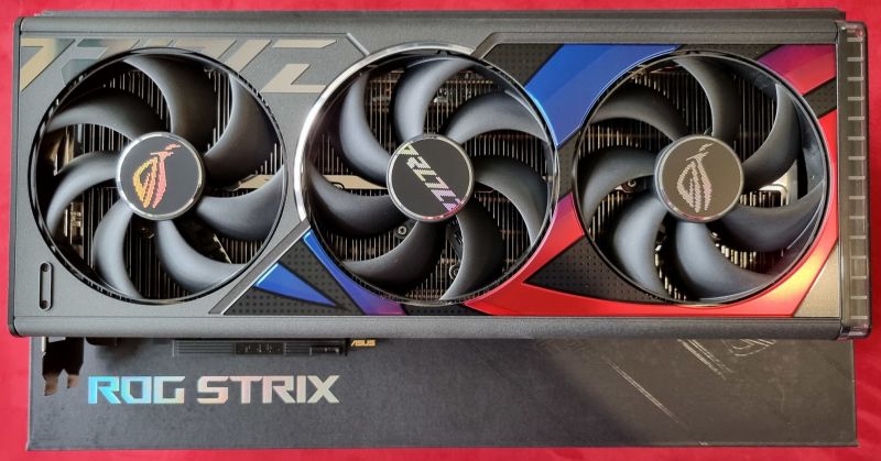 Vue de face d'une carte graphique noire à trois grands ventilateurs, avec des inserts bleus et rouges, posée sur sa boîte marquée « ROG Strix » devant un fond rouge.