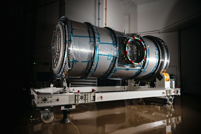 Un long cylindre métallique argenté, cerclé de bandes bleues, avec une écoutille ronde sur le côté, posé sur un châssis de transport blanc à roulettes dans un hall sombre au sol brillant.