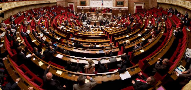 Vue panoramique de l'hémicycle du Palais-Bourbon en séance : des députés assis sur les bancs de bois aux fauteuils rouges, disposés en demi-cercle face à la tribune et au perchoir.