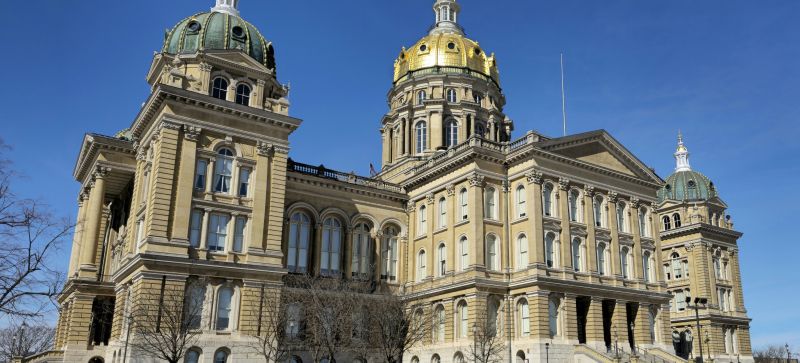 Le Capitole de l'Iowa, grand bâtiment de pierre claire surmonté d'un dôme doré et flanqué de dômes verts, sous un ciel bleu, derrière des arbres sans feuilles.