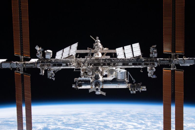 La Station spatiale internationale en orbite, vue de face sur fond noir : sa longue poutre centrale, les modules pressurisés au centre, des radiateurs blancs et, aux deux extrémités, de grands panneaux solaires orangés ; la courbure de la Terre, couverte de nuages, occupe le bas de l'image.