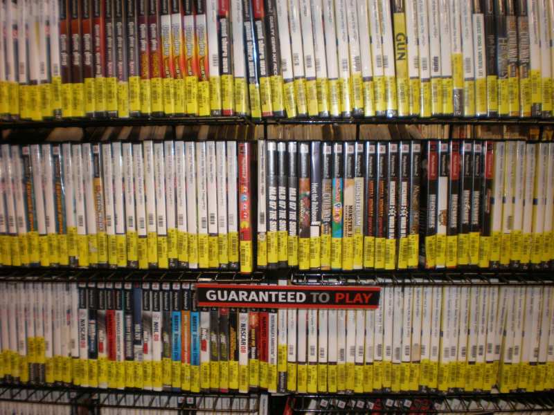 Rayonnages métalliques d'un magasin garnis de boîtiers de jeux PlayStation 2 d'occasion, étiquettes de prix jaunes au pied de chaque tranche, et un panneau « Guaranteed to play ».