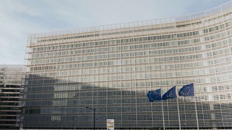 La façade claire et incurvée d'un grand immeuble de bureaux, couverte de lamelles horizontales, sous un ciel voilé ; trois drapeaux européens bleus étoilés flottent devant le bâtiment.