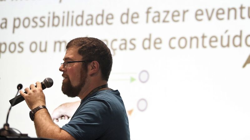 Un homme barbu à lunettes, en tee-shirt bleu, parle dans un micro devant un grand écran de projection couvert d'un texte en portugais.
