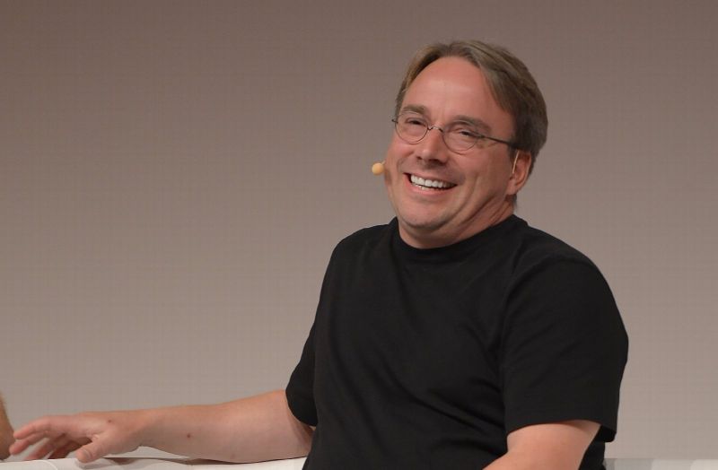 Linus Torvalds, assis sur un canapé blanc, sourit sur une scène de conférence, un micro-casque à l'oreille.