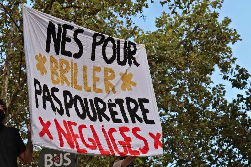 Une banderole blanche tenue à bout de bras sous des platanes, sur laquelle est peint « Nés pour briller, pas pour être négligés ».