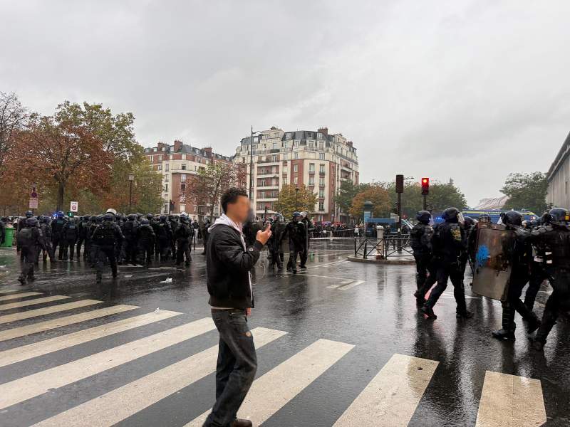 Sur un passage piéton mouillé par la pluie, un homme au visage flouté tient un téléphone devant lui et en regarde l'écran ; autour de lui, des colonnes de membres des forces de l'ordre casqués, avec boucliers, devant des immeubles et un fourgon de gendarmerie.