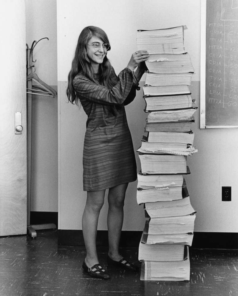 Photo en noir et blanc : une jeune femme aux longs cheveux et aux lunettes rondes, en robe rayée, sourit debout à côté d'une pile de classeurs de listings imprimés aussi haute qu'elle, dans un bureau avec un tableau noir couvert d'inscriptions.