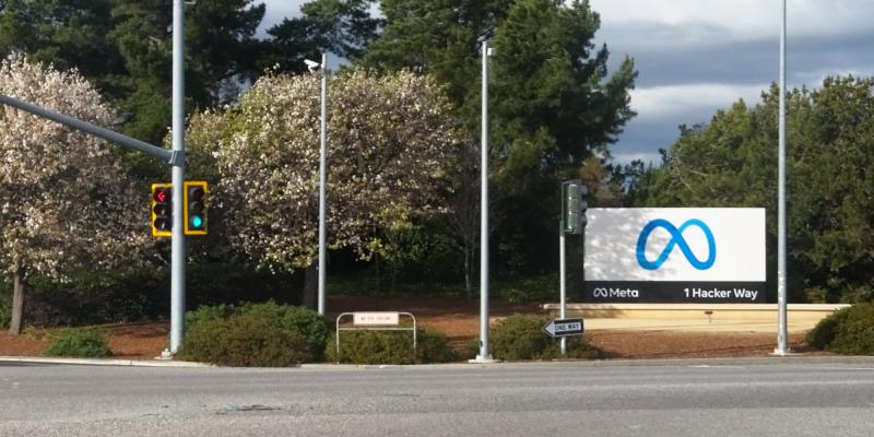 Au bord d'un carrefour, entre des arbres en fleurs et des feux tricolores, un panneau blanc porte le logo bleu de Meta et l'adresse 1 Hacker Way.