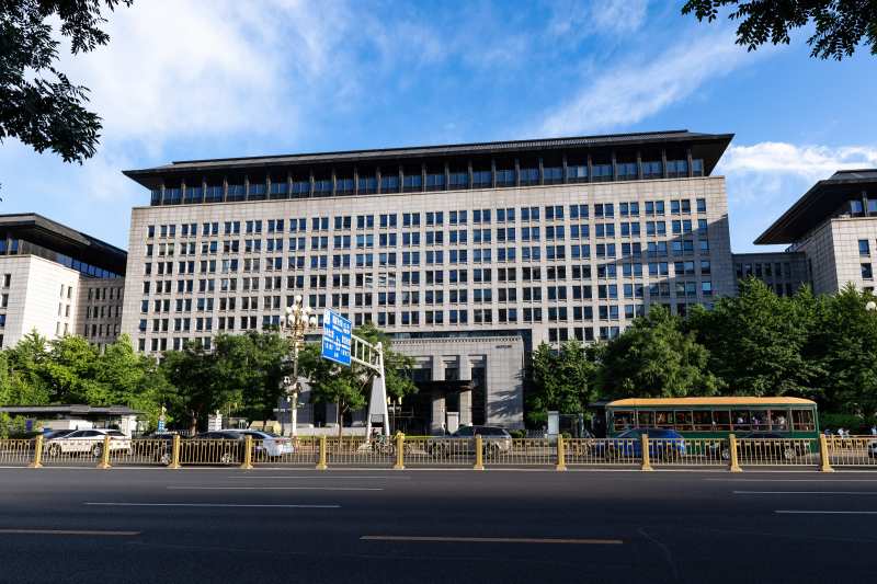 Grand immeuble de bureaux gris du ministère chinois du Commerce, à Pékin, vu depuis une avenue bordée d'arbres, sous un ciel bleu