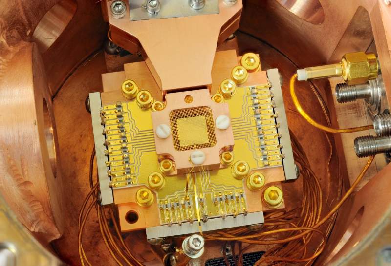 Puce carrée d'un piège à ions, couverte d'une grille dorée, au centre d'un support aux pistes et vis dorées, dans une enceinte en cuivre