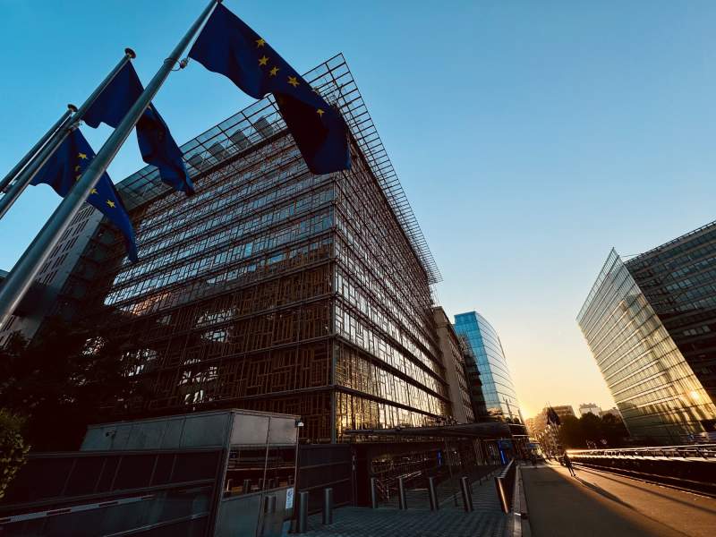 Le bâtiment Europa à Bruxelles, vu en contre-plongée : façade en mosaïque de vieilles fenêtres derrière une résille de verre, drapeaux européens au premier plan, rue éclairée par le soleil couchant.