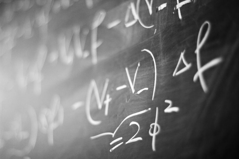 Gros plan en noir et blanc, pris de biais, sur un tableau noir couvert d'équations écrites à la craie : au premier plan, nets, « = 2φ² » et « Δf » ; le reste des formules se perd dans le flou.