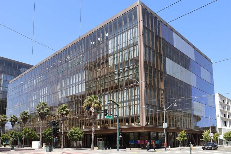 Grand immeuble de bureaux cubique aux façades de verre et à l'armature cuivrée, à un angle de rue planté de palmiers, sous un ciel bleu traversé par des câbles aériens.