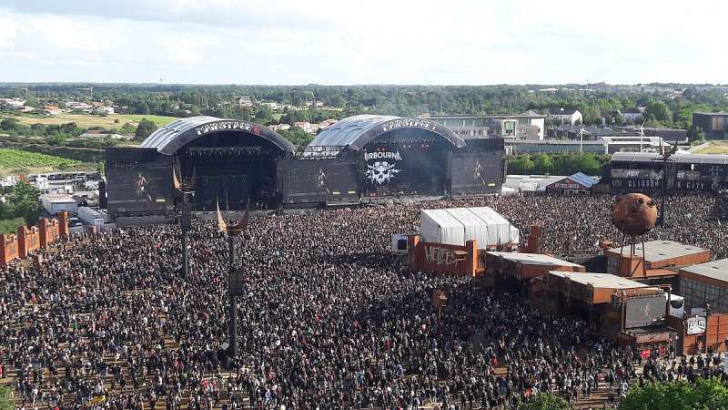 Vue plongeante, en plein jour, sur une foule compacte de festivaliers massés devant deux grandes scènes couvertes, avec la campagne et des habitations en arrière-plan.
