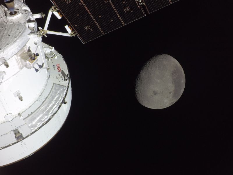 Au premier plan, une partie du vaisseau Orion éclairée par le Soleil ; derrière lui, sur fond noir, la Lune presque pleine, avec en bas un grand bassin circulaire cerné de reliefs.
