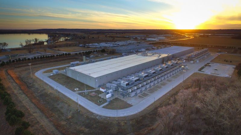 Vue aérienne, au coucher du soleil, d'un centre de données de Google : un long bâtiment bas au toit blanc, bordé de rangées d'équipements techniques, près d'un plan d'eau.