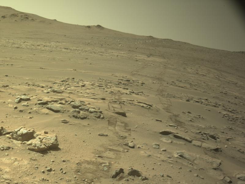 Une pente de sable ocre parsemée de pierres plates et de blocs clairs ; deux paires de traces de roues striées partent du bas de l'image et remontent vers une crête, sous un ciel jaune pâle.