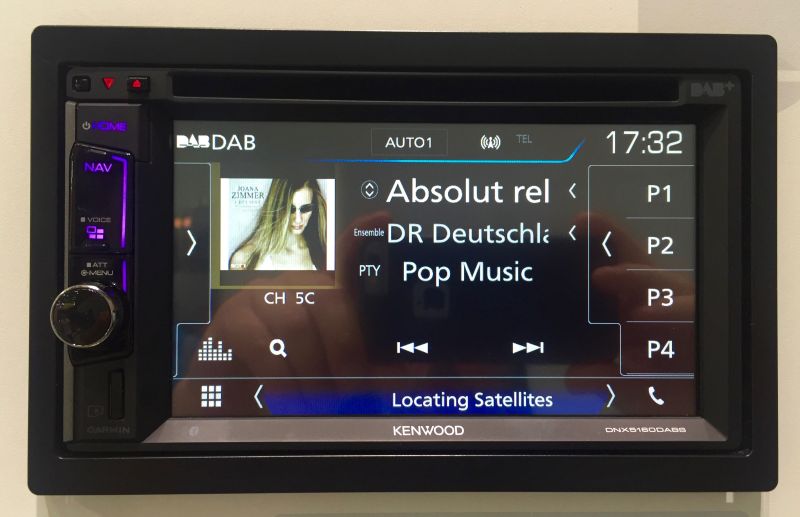 Autoradio Kenwood à écran tactile fixé sur un présentoir, l'écran en mode DAB affichant le nom d'une station allemande et la pochette du titre diffusé.
