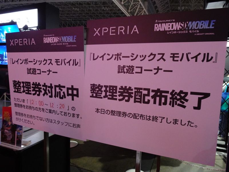 Deux panneaux roses imprimés en japonais, frappés en tête des logos Xperia et Rainbow Six Mobile, posés à l'entrée d'un espace d'essai d'un salon de jeux vidéo.