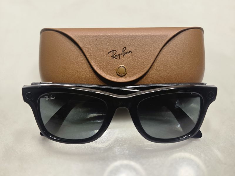 Paire de Ray-Ban Meta noires et brillantes, de première génération, posée devant son étui de recharge en cuir marron.