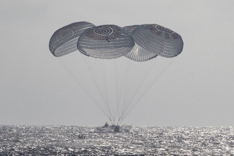 Quatre grands parachutes blanc et rouille, réunis au-dessus de la mer, retiennent par de longues suspentes une capsule qui touche l'eau dans une gerbe d'écume ; la mer scintille sous un ciel gris et voilé.