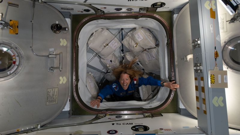 Vue de dessus d'une écoutille carrée ouverte, encadrée de deux panneaux métalliques ; une astronaute en combinaison bleue, cheveux flottant en apesanteur, la franchit bras écartés et bouche ouverte, devant des sacs de transport blancs.
