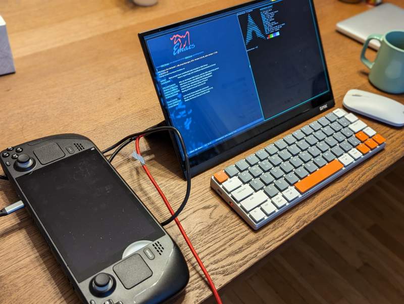 Une console Steam Deck posée sur un bureau en bois, reliée par câble à un écran portable qui affiche l'éditeur Emacs et un terminal avec le logo d'Arch Linux, devant un clavier compact.