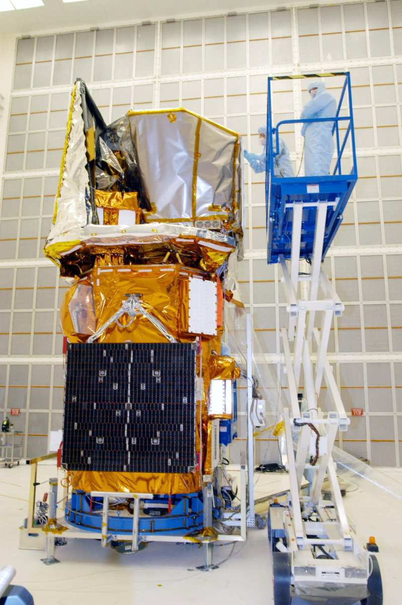 Un gros satellite enveloppé de couvertures thermiques dorées et argentées, dans un hangar aux murs blancs ; à droite, sur une nacelle élévatrice, deux techniciens en combinaison de salle blanche bleu clair travaillent sur sa partie haute.