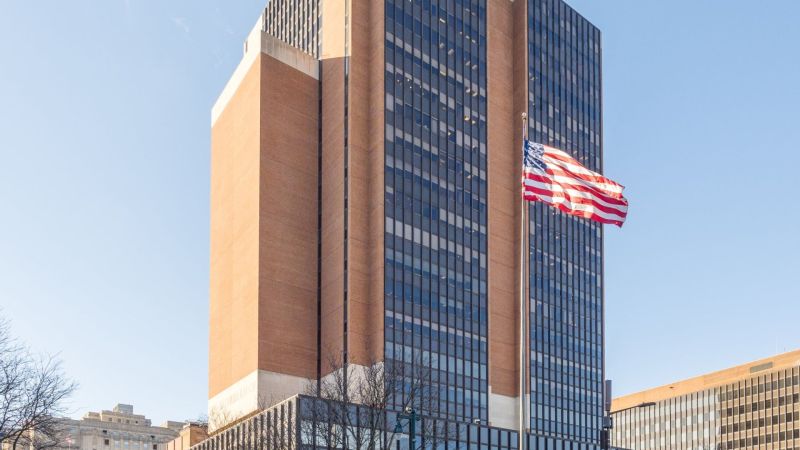 Une tour de bureaux aux façades de brique et de verre sombre se dresse sous un ciel bleu ; au premier plan flotte un drapeau américain.