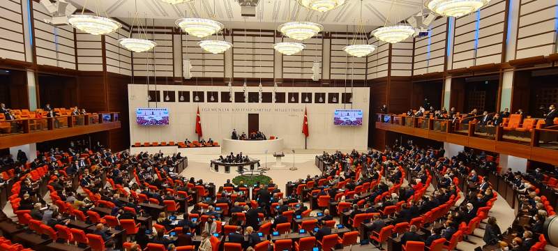 Vue panoramique de l'hémicycle de la Grande Assemblée nationale de Turquie en séance : des centaines de députés assis sur des fauteuils orange, sous de grands lustres, face à la tribune encadrée de deux drapeaux turcs ; au mur, l'inscription « Egemenlik kayıtsız şartsız milletindir ».