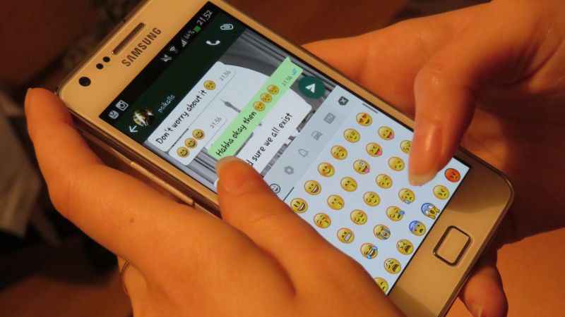 Deux mains tiennent un smartphone Samsung blanc ouvert sur une conversation WhatsApp, clavier d'émojis affiché.