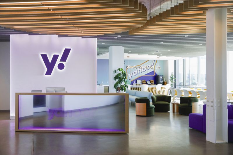 Accueil des bureaux de Yahoo : comptoir vitré devant un mur blanc portant le « y! » violet lumineux, salon et cafétéria au fond.